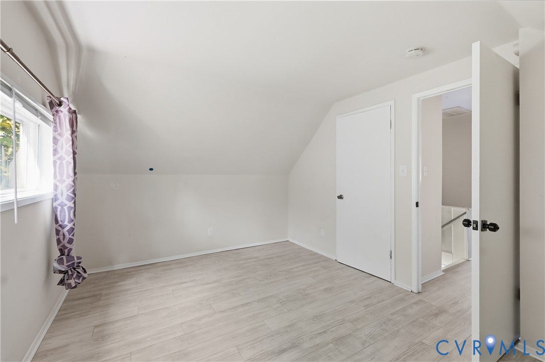 Vaulted ceiling, light wood-finish flooring, white trim, interior doors with dark hardware, and a window with exterior visibility