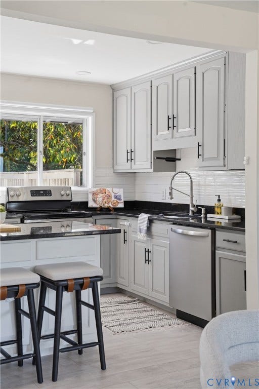 Kitchen featuring light gray cabinetry, dark countertops, a tile backsplash, stainless steel appliances, and light wood-finish flooring