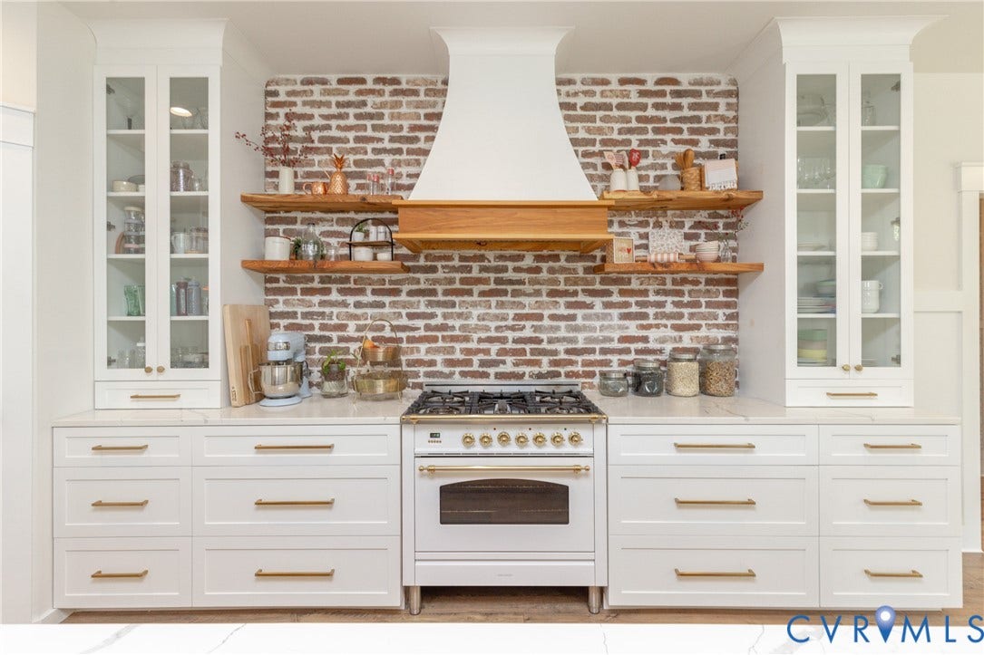 White cabinetry with gold-tone hardware, exposed brick backsplash, wood floating shelves, white range hood, and a white range with gold-tone accents