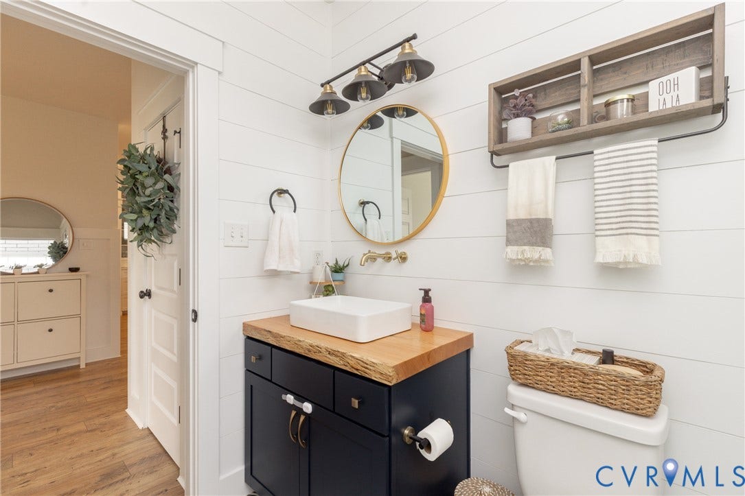 Shiplap accent wall, dark-toned vanity with wood-finish countertop, vessel sink, gold-toned wall-mounted faucet, and a round gold-framed mirror