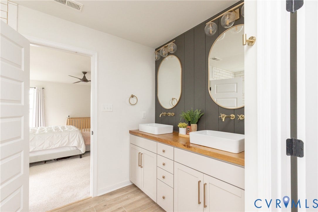 Dual vessel sinks with wall-mounted faucets, oval mirrors, wood-finish countertop, and white cabinetry with gold-tone hardware