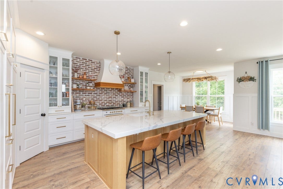 Spacious kitchen featuring an expansive island with a stone-finish countertop, white cabinetry with gold-toned hardware, a brick-finish backsplash, and wood-finish flooring