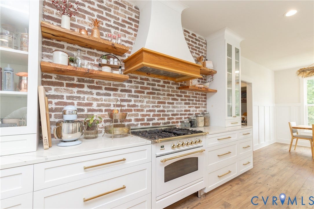 White cabinetry with gold-tone hardware, wood-finish flooring, brick-finish backsplash, and an integrated range with a custom wood and plaster hood