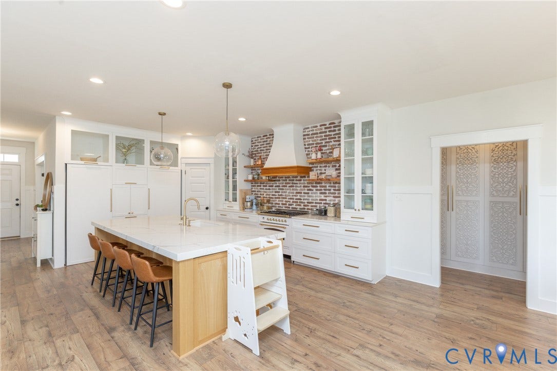 Spacious kitchen featuring an expansive island with seating, wood-finish flooring, white cabinetry with gold hardware, and a brick-look backsplash