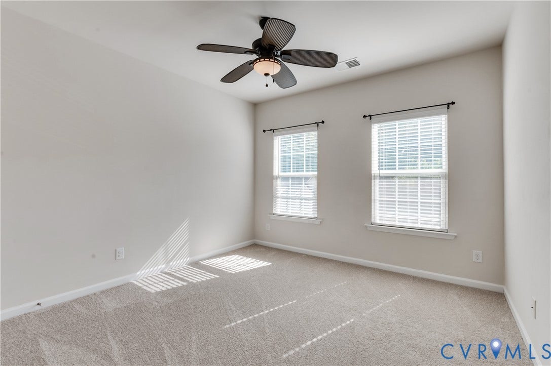 Bright, light-filled bedroom featuring dual windows with plantation shutters, neutral carpeting, ceiling fan with integrated lighting, and fresh white walls