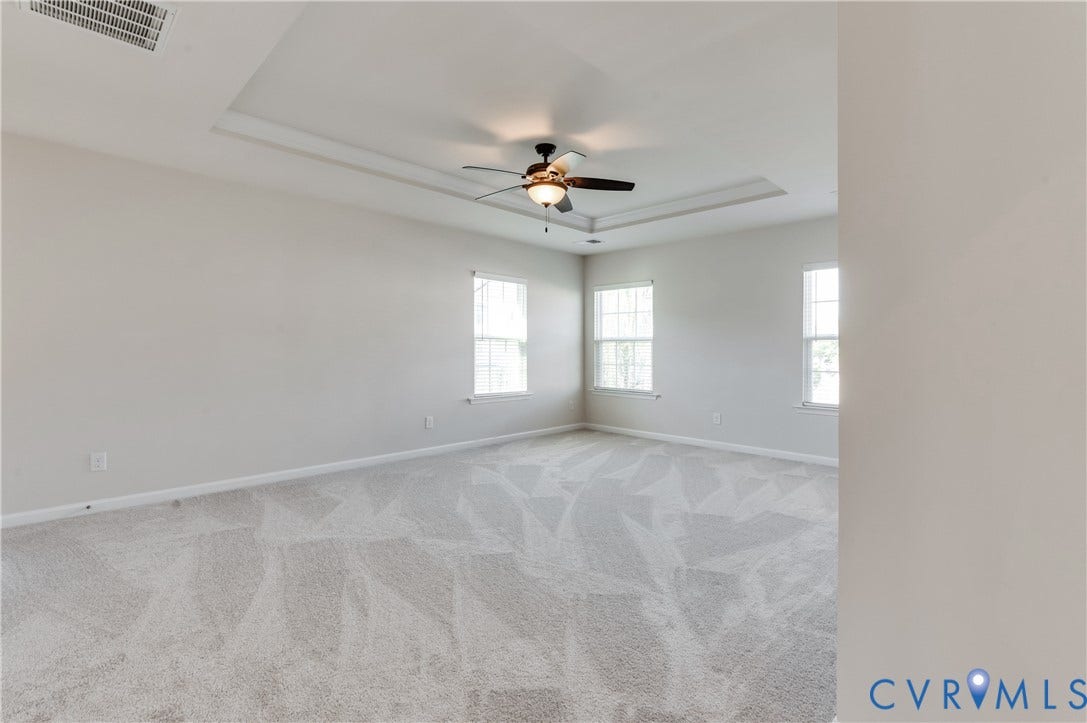 Recessed tray ceiling with a ceiling fan, light gray carpeting, and multiple windows