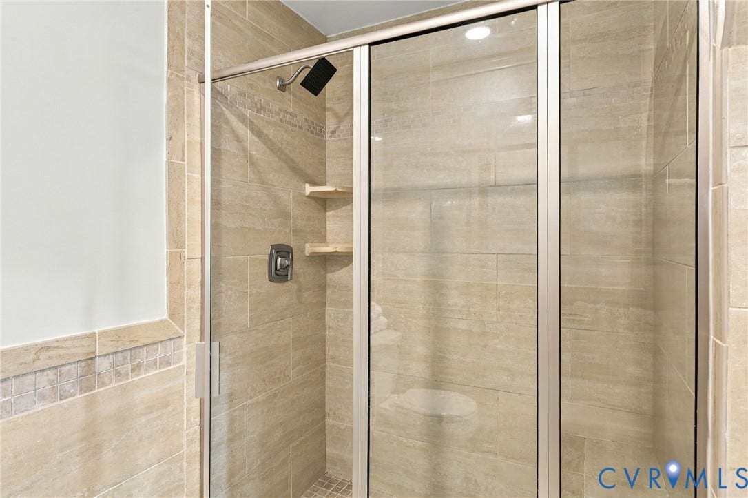 6409 Walmsley Boulevard, Chesterfield, VA 23224 Walk-in shower featuring floor-to-ceiling neutral tile, semi-frameless glass enclosure, matte black rainfall showerhead, integrated corner shelving, and mosaic tile accents