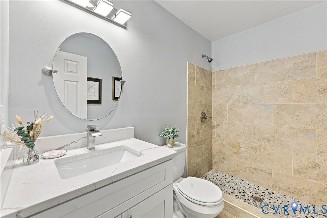 6409 Walmsley Boulevard, Chesterfield, VA 23224 Bathroom vanity with white stone-finish countertop, undermount sink, and oval mirror, accompanied by a walk-in shower featuring textured stone tile walls and a pebble-stone floor