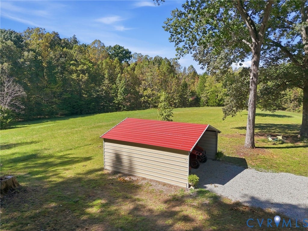 Detached carport with a red metal roof, gravel driveway, and expansive grassed area bordered by mature trees.
