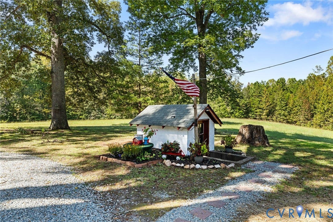 Gravel driveway with stepping stones, bordered by a small structure with a gabled roof, surrounded by lawn and mature trees.