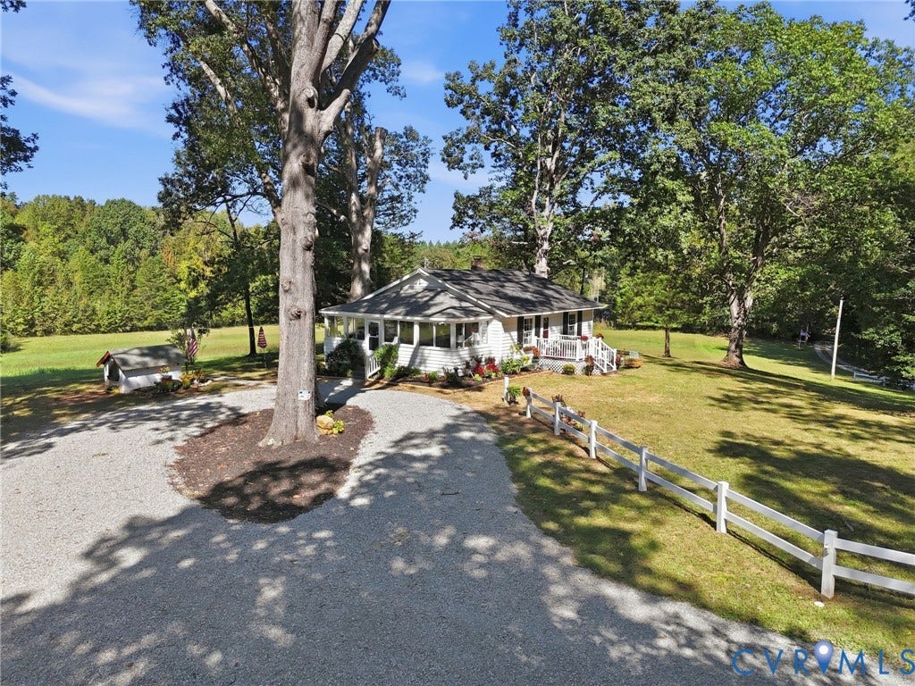 Gravel driveway with mature trees, white picket fence, and a sunroom with multiple windows.