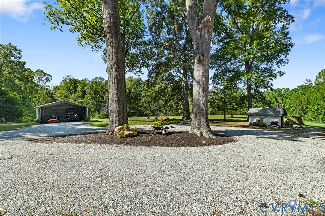 Gravel driveway with mature trees, open-sided metal structure, and a secondary small outbuilding.