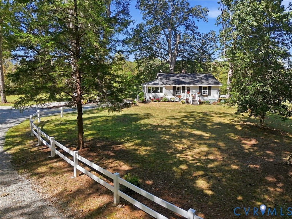 White picket fence delineating the front yard, mature trees providing natural shading, gravel driveway, and a single-story residence with dark shutters.