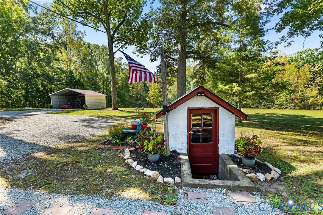 Gravel driveway with a metal carport, a small outbuilding featuring a red entry door, and a landscaped garden bed.