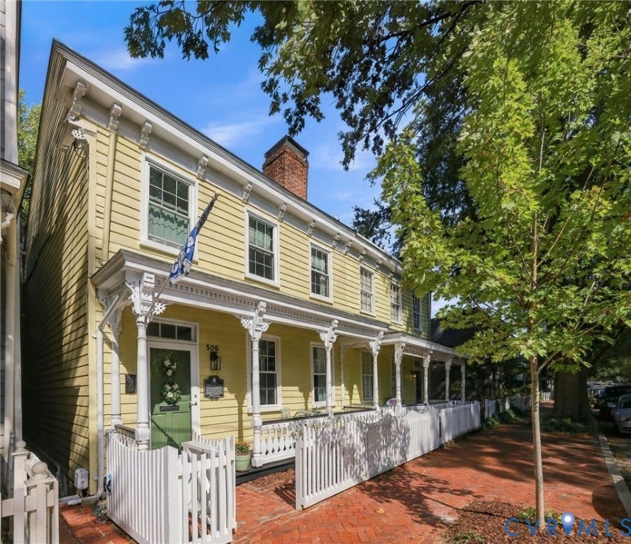 Yellow siding exterior with white trim, brick chimney, covered porch with decorative pillars, and white picket fence