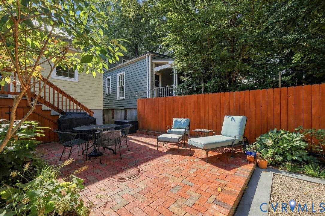 Brick paver patio, wood privacy fence, gravel border, mature greenery, and exterior stairs with wood railing