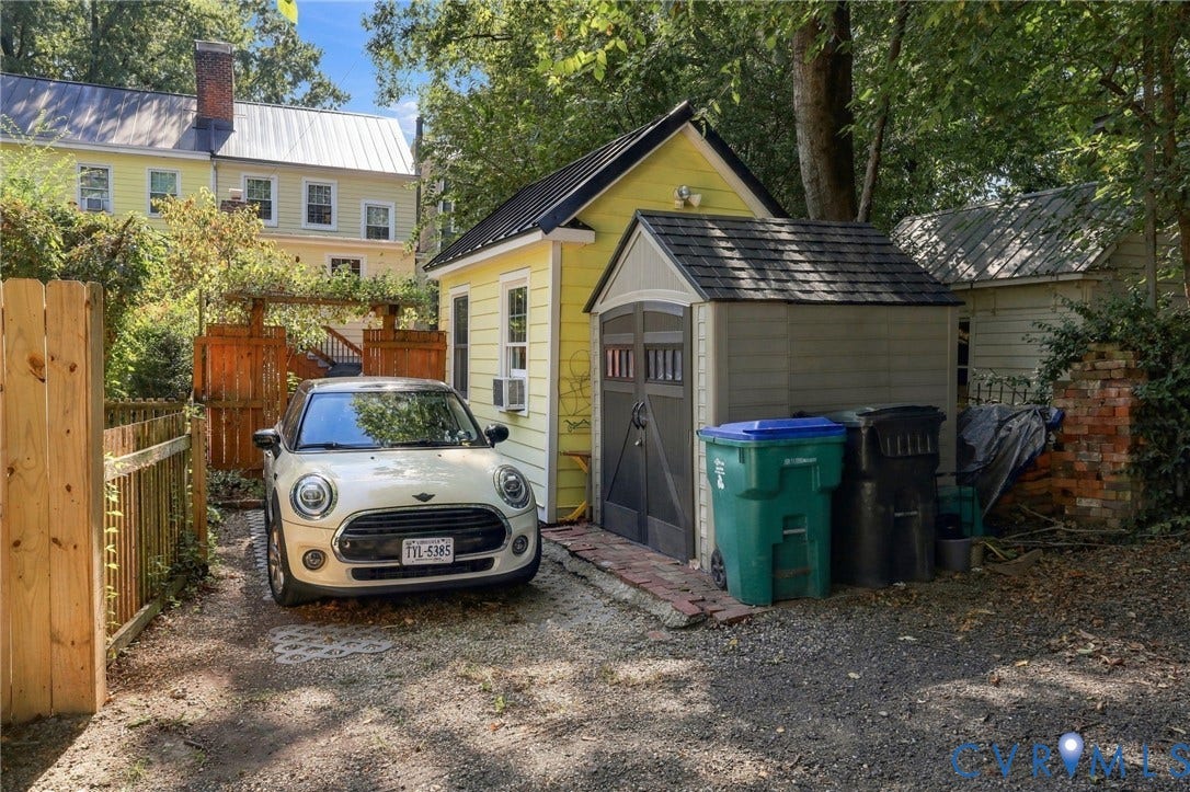 Gravel driveway with brick trim, wood fencing, and a detached structure featuring yellow siding and a black metal roof
