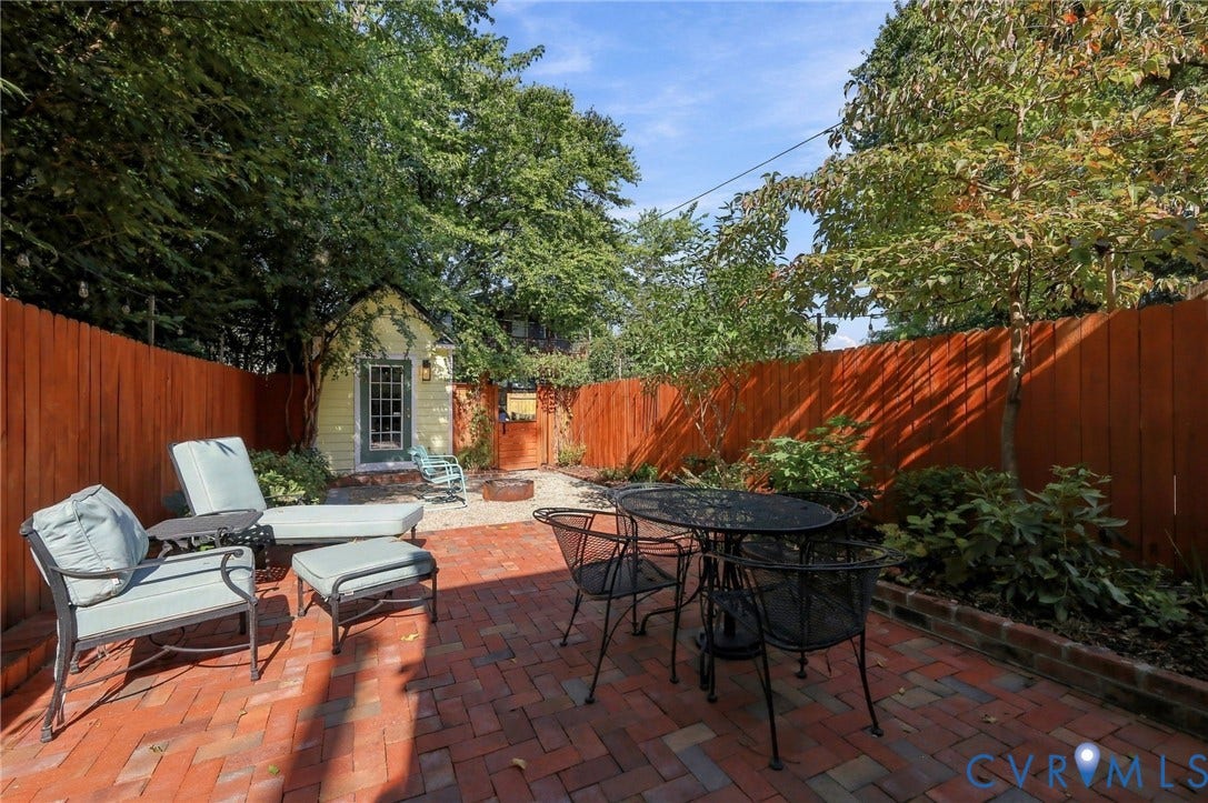 Paved brick patio with an enclosed wood fence and a separate yellow outbuilding, complemented by mature trees and landscaping