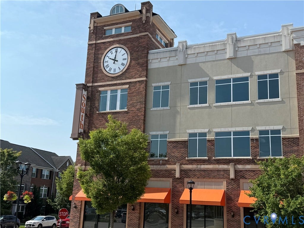 3931 Liesfeld Place, West Broad Village, Henrico, VA 23060 Historic brick mixed-use building featuring a prominent clock tower with illuminated dial, large storefront windows with orange awnings on the ground level, and a contemporary tan faade addition
