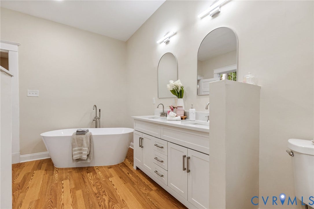 12105 Mathenay Drive, Mathenay, Chesterfield, VA 23831 White freestanding bathtub with a floor-mounted faucet, dual arched vanity mirrors, double sink vanity with white cabinetry, and wood-finish flooring