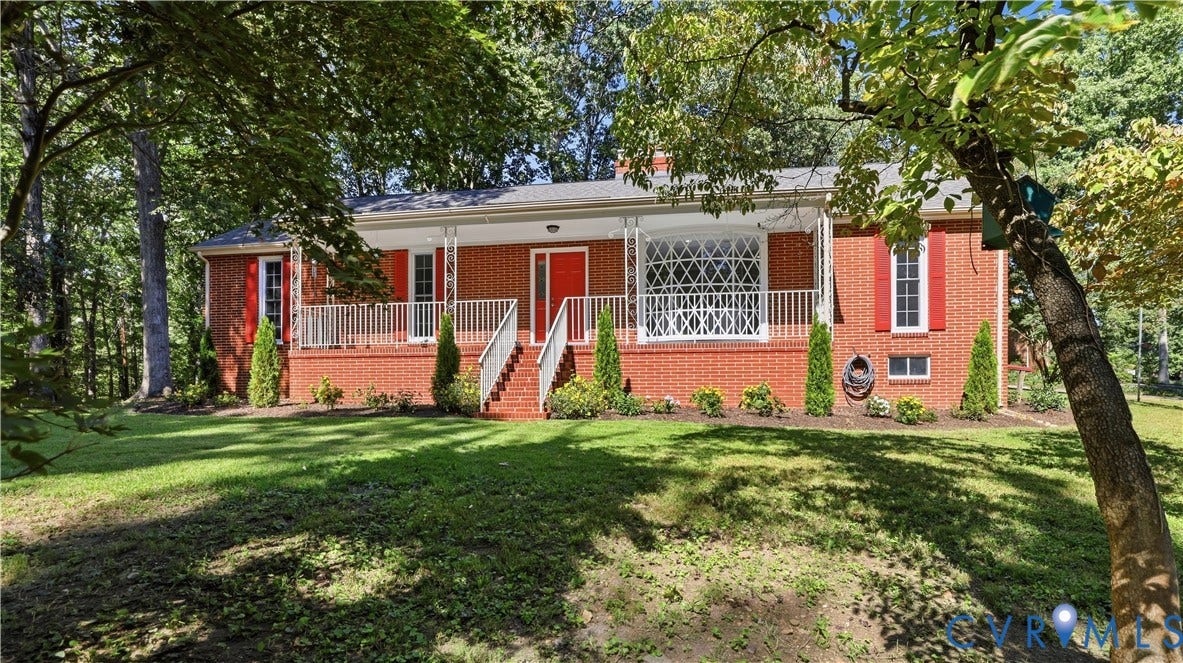 Brick exterior with red shutters, a covered front porch, white railings, and mature trees