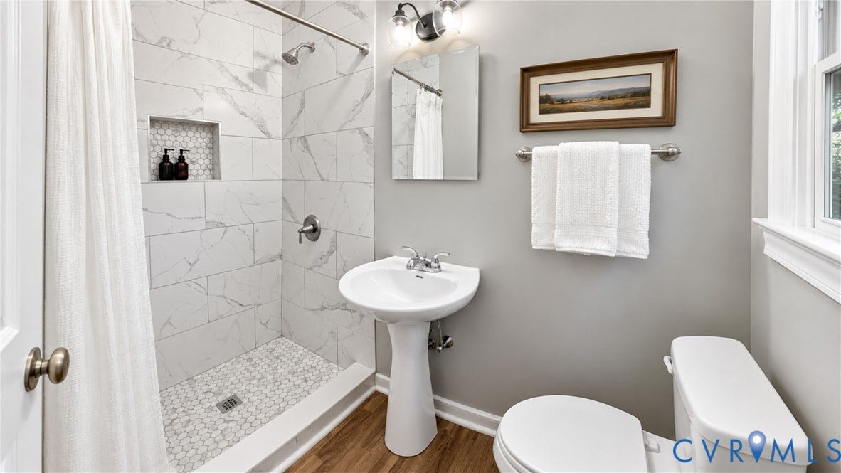 Shower with white subway tile surround and mosaic tile floor, pedestal sink, and wood-finish flooring
