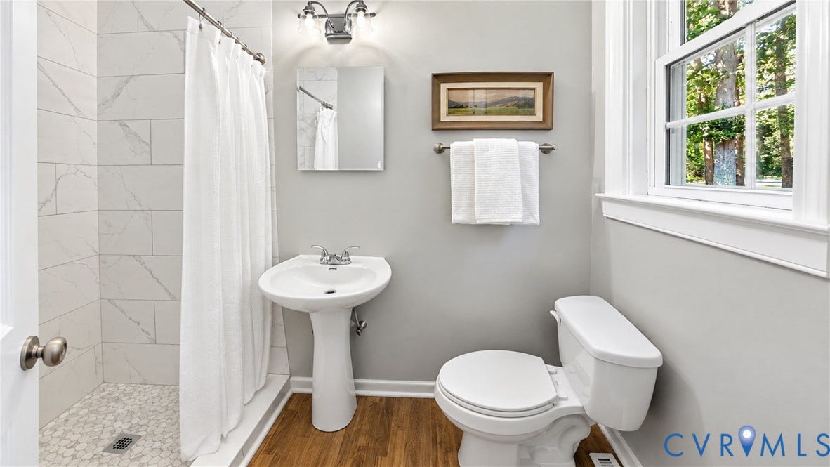 Pedestal sink, wood-finish flooring, tiled shower with pebble floor, window with natural light, and contemporary vanity lighting