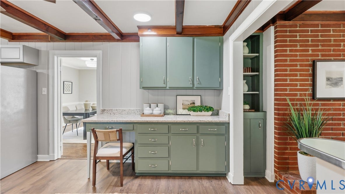 Exposed wood ceiling beams, green cabinetry with ample storage, light-toned countertop, wood-finish flooring, and a red brick wall accent