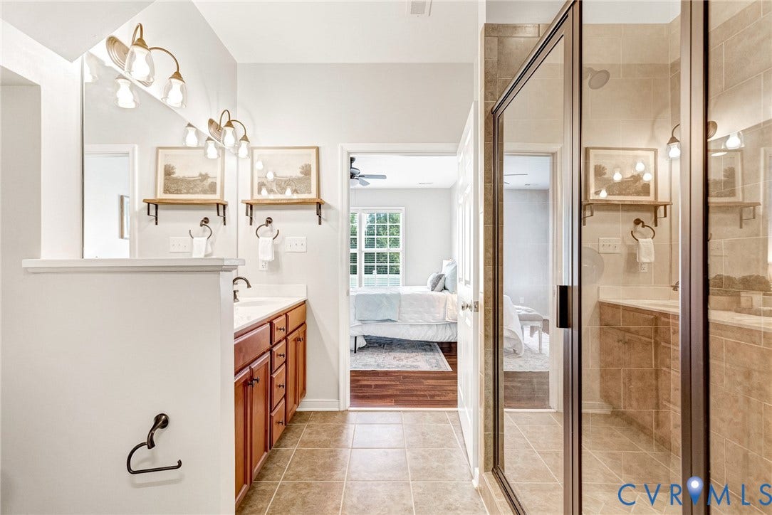 7728 Marshall Arch Drive, Liberty Trace, Mechanicsville, VA 23111 Dual vanity with wood cabinetry and undermount sinks, bronze-finish wall sconces, large wall-mounted mirror, and a tiled walk-in shower with a glass enclosure