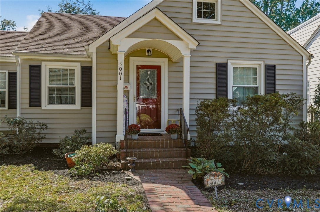 Gabled roofline, light-toned siding, red paneled front door with decorative glass, white-trimmed windows with dark shutters, and a brick walkway leading to the entrance
