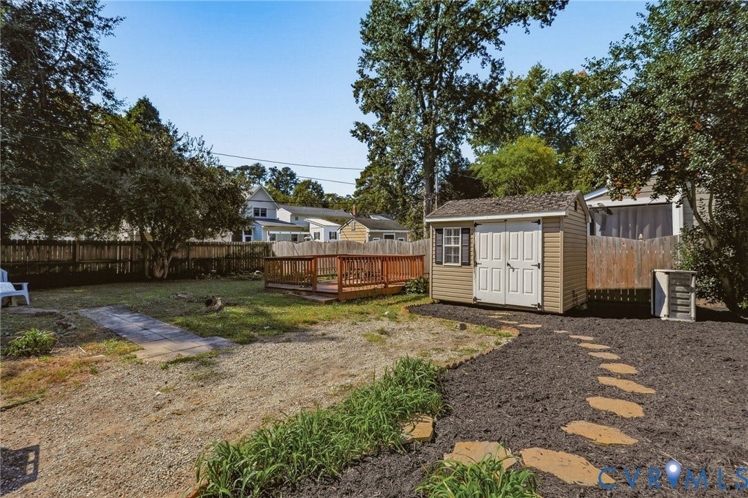 Expansive backyard featuring a wood deck, a storage shed with double doors, stone pathway, and mature perimeter trees