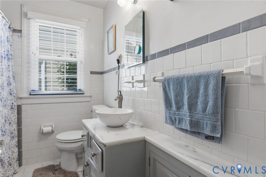 White subway tile walls with a gray trim accent, a window with horizontal blinds, a white vessel sink, a light gray vanity with drawer and cabinet storage, and a white countertop