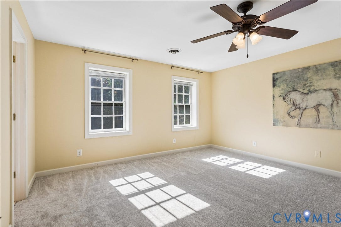 Spacious room featuring light-colored walls, light grey carpet, two double-hung windows, and a ceiling fan with integrated lighting