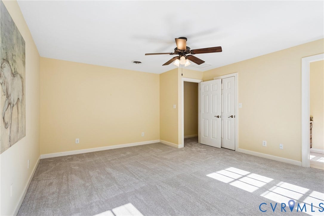 Spacious room with light-toned walls, grey carpeting, and a ceiling fan with light fixture