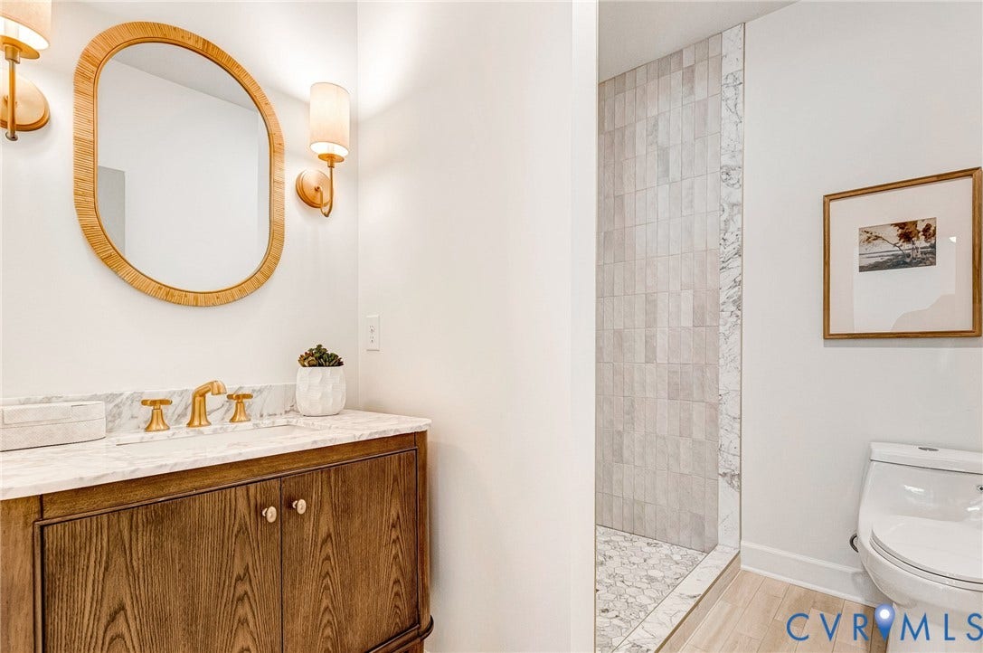 404 Waveny Road, Westham, Henrico, VA 23229 Wood-finish cabinetry featuring brass hardware, stone-top vanity with widespread faucet, oval mirror with textured frame, dual wall-mounted light fixtures, and walk-in shower with vertical tile surround