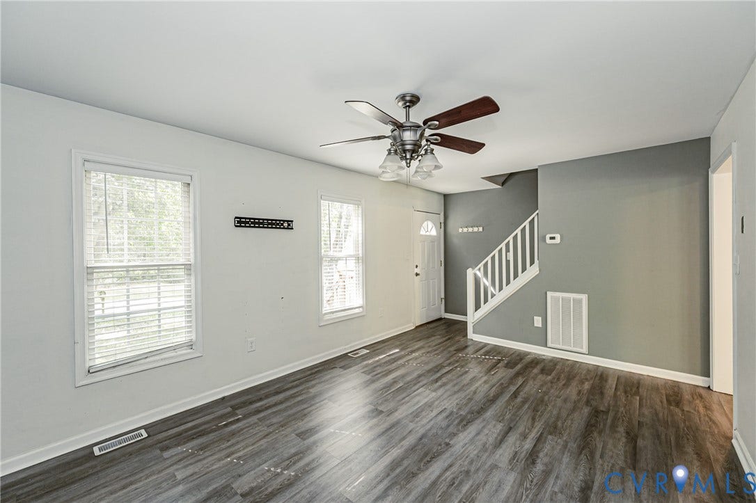 Spacious open living area featuring wood-finish flooring, abundant natural light from dual windows, ceiling fan with integrated lighting, and a staircase with white railings leading to upper level