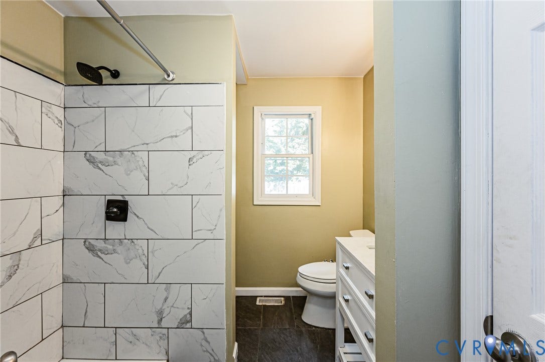 Shower with white subway tile and dark fixtures, dark tiled flooring, white vanity with white countertop, and a window