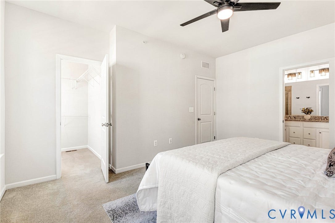 Neutral tone wall paint, ceiling-mounted fan, carpeted flooring, walk-in closet with wire shelving, and an en-suite bathroom with a double vanity featuring a stone countertop