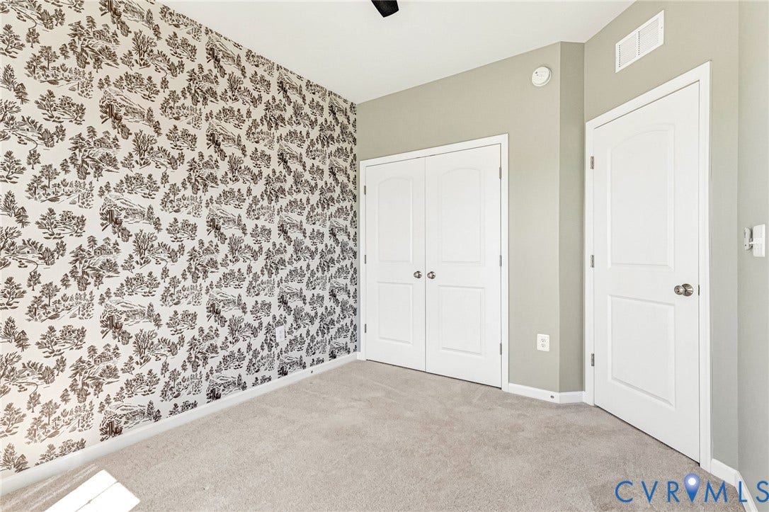 Patterned accent wall, light grey wall paint, light grey carpeting, white paneled doors, and ceiling fan