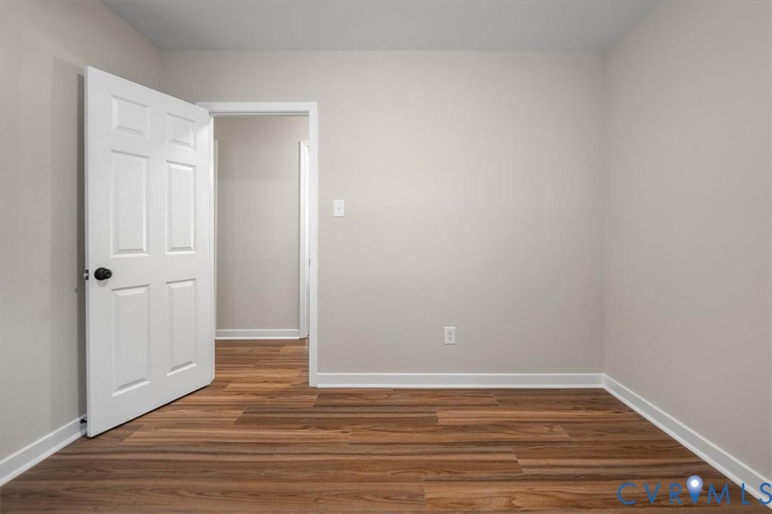 Wood-finish flooring throughout, light neutral wall paint, white trim, and a white six-panel door with black hardware