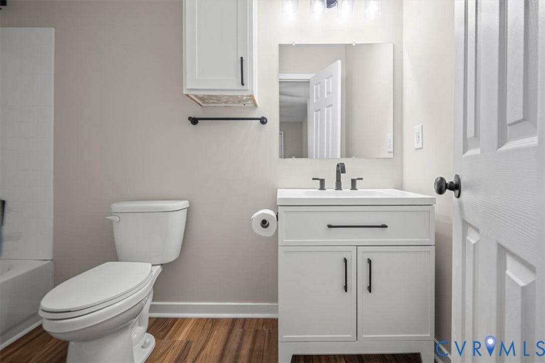White vanity with matte black hardware, wood-finish flooring, wall-mounted mirror, and overhead cabinet storage