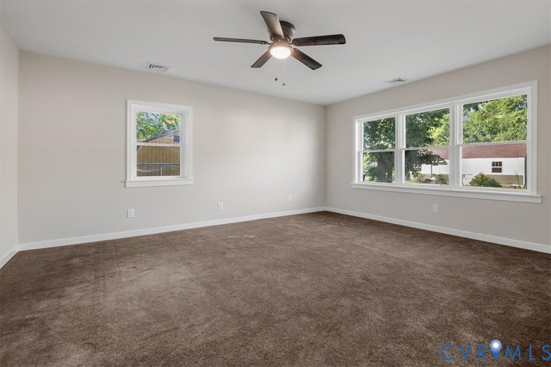 Spacious room featuring neutral wall tones, brown carpeting, multiple windows providing natural light, white trim, and a ceiling fan with integrated lighting