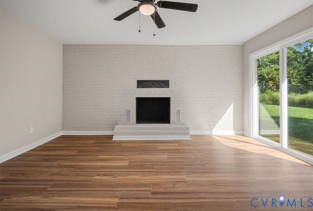 Wood-finish flooring, painted brick fireplace with hearth, ceiling fan with integrated lighting, and white-framed sliding glass doors