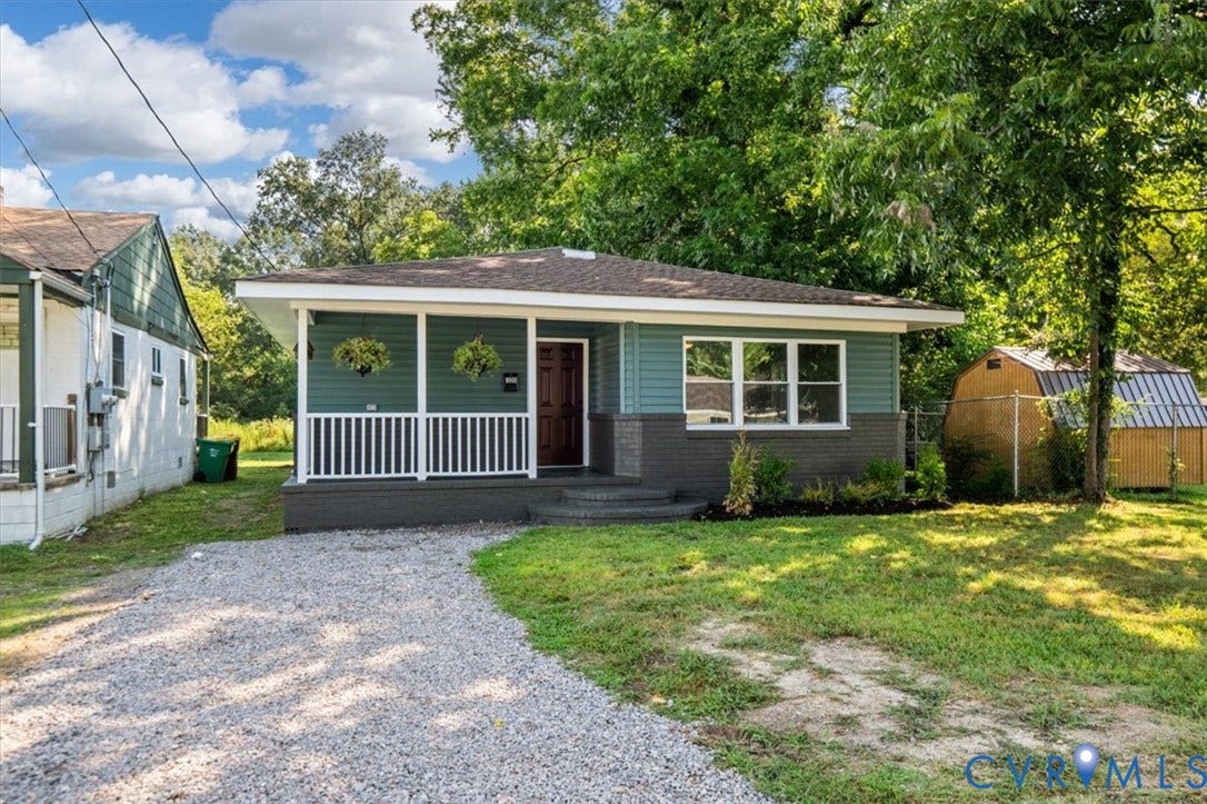 Property featuring a covered porch with white railing, a gravel driveway, mature trees, and an accessory structure