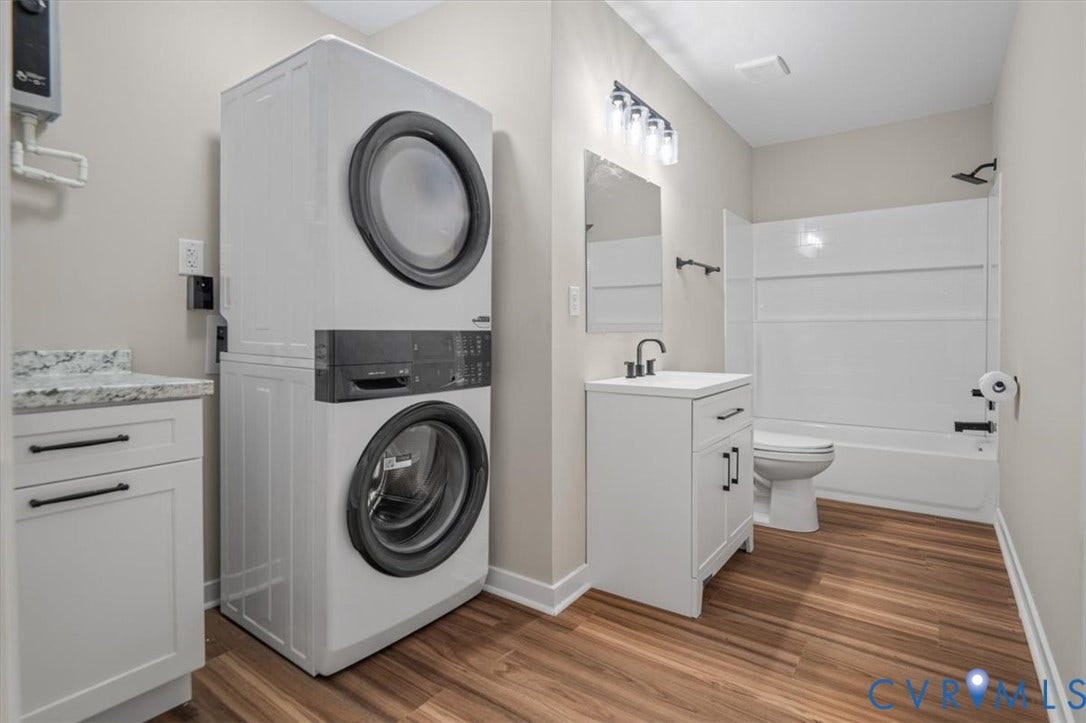 Wood-finish flooring, white vanity with matte black faucet, full bathtub with shower, stackable washer and dryer, and white cabinetry with granite-style countertop