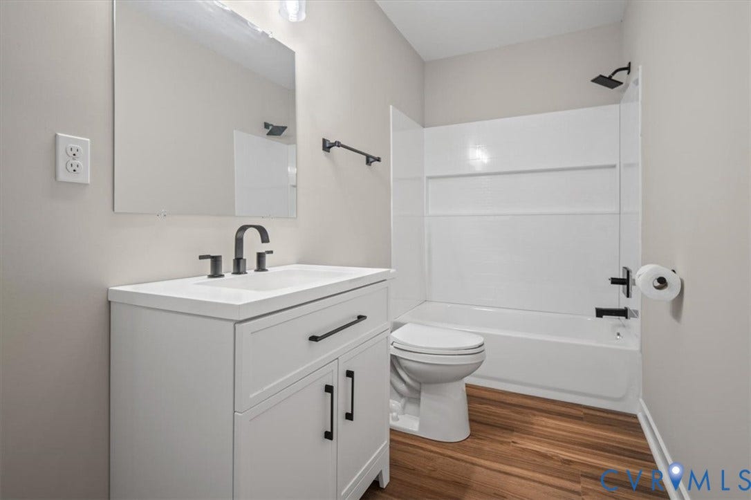 White vanity with a single basin sink and matte black hardware, white bathtub with a shower kit, and wood-finish flooring