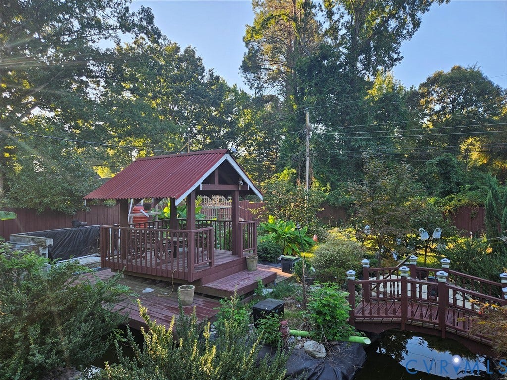 Expansive outdoor living area featuring a gazebo with a red corrugated roof, a wooden deck, a decorative pond with a footbridge, and mature landscaping