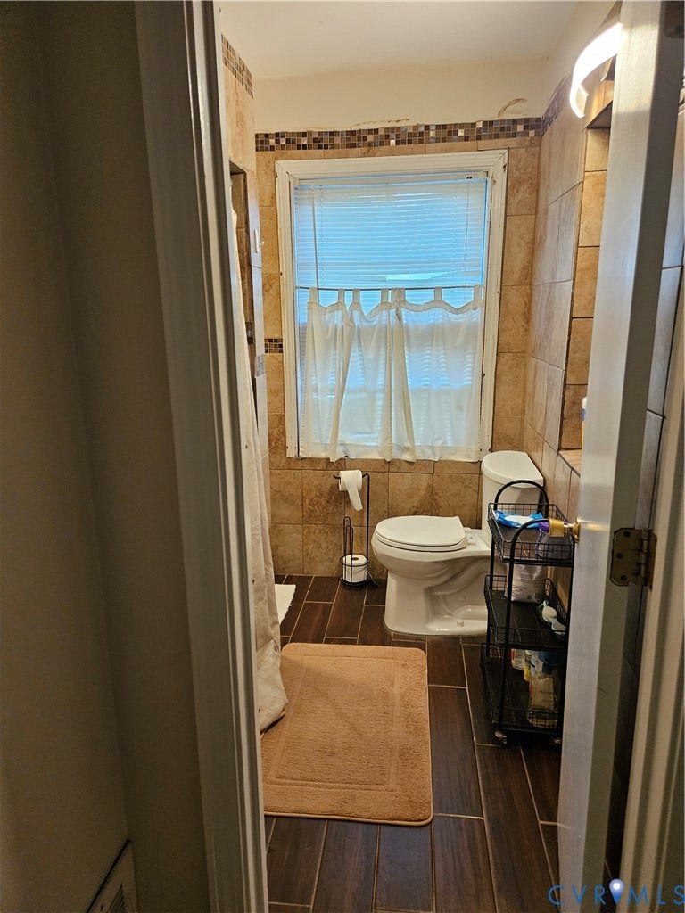 Tile flooring with a wood-finish, ceramic wall tiling, mosaic tile accent, and a single window with blinds