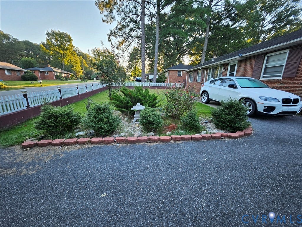 Paved driveway, brick exterior, white picket fence, landscaped front yard with decorative stone accents, and mature trees