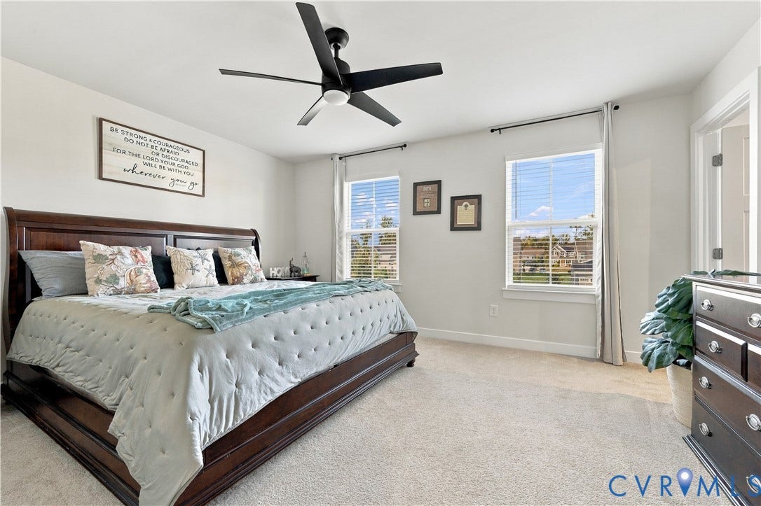 Spacious room featuring a dark-toned ceiling fan with integrated lighting, two windows with blinds, light-toned carpeting, and light-colored walls